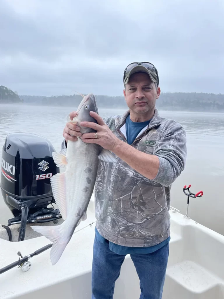 A man in a camo jacket and cap holds a large fish on a boat with a Suzuki boat motor, against a backdrop of calm, misty water, reminiscent of early mornings spent picking inventory in a quiet warehouse.