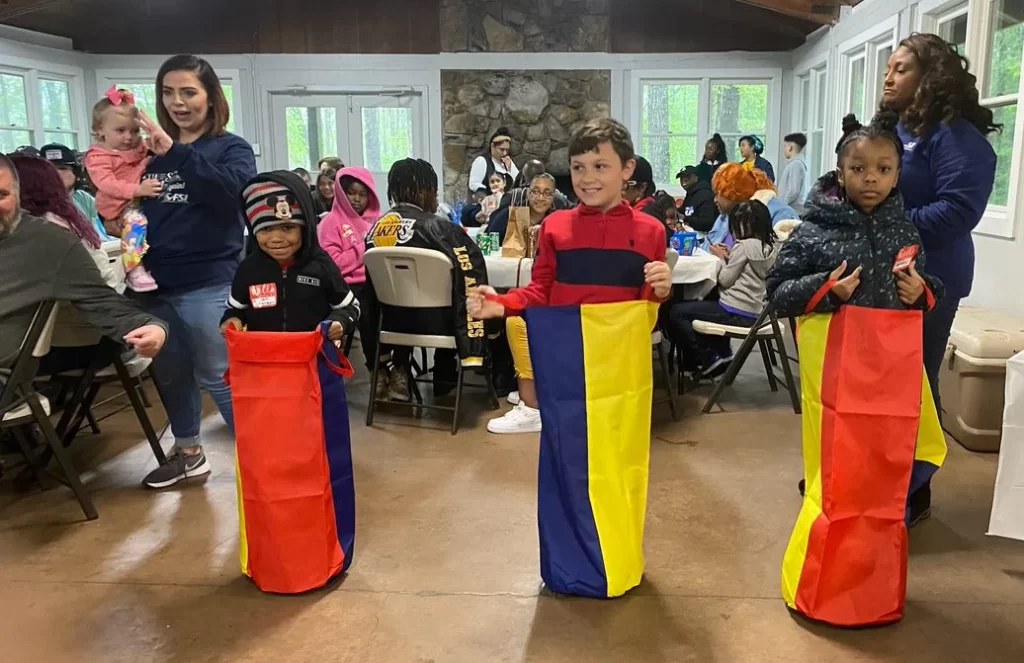 Children participate in sack races indoors while adults and other children watch and engage in activities, such as organizing shipping boxes at the tables in the background.