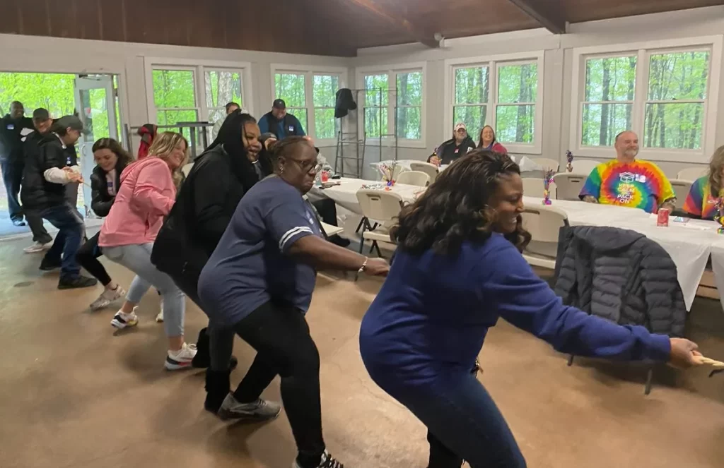 A group of people participates in a line dance inside a spacious warehouse with tables and chairs, while others sit and watch.