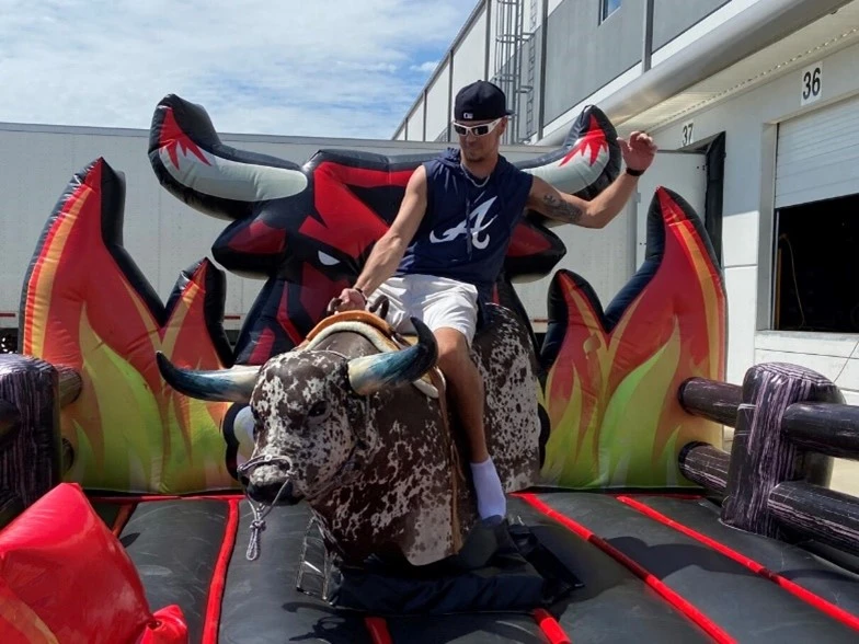A person rides a mechanical bull in an inflatable arena with red and yellow flame designs, resembling the hustle of picking boxes in a busy warehouse. The rider is wearing a sleeveless shirt, shorts, cap, and sunglasses.