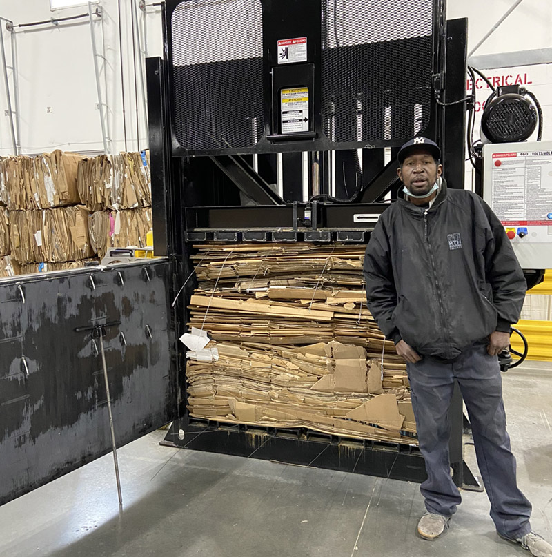 A man stands next to a cardboard baler machine with compressed cardboard boxes inside it, in a bustling warehouse setting. Stacks of flattened cardboard are visible in the background, highlighting the efficiency of logistics and fulfillment operations.