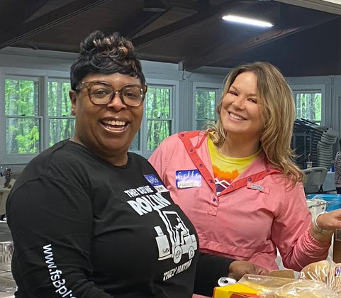 Two women are smiling while standing together indoors. One wears a black long-sleeve shirt, and the other is in a pink jacket. Both appear happy and are surrounded by boxes in a room with large windows, likely discussing logistics for their latest project.