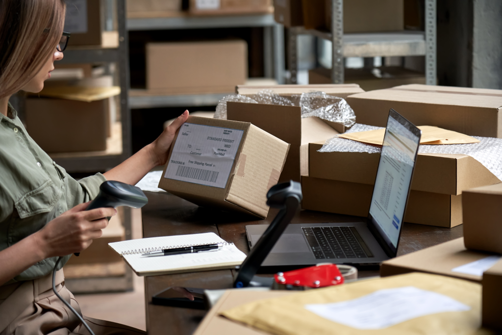 A woman holding a package and scanning its barcode at a workstation with a laptop, various boxes, and shipping supplies in a bustling warehouse setting.