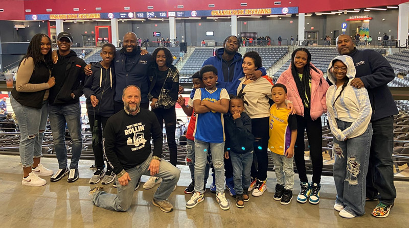 A group of people, including adults and children, are posing for a photo in an indoor stadium setting. Some are standing, while a few kneel amidst stacks of boxes. The stadium seating is visible in the background, giving the impression of a busy warehouse atmosphere.